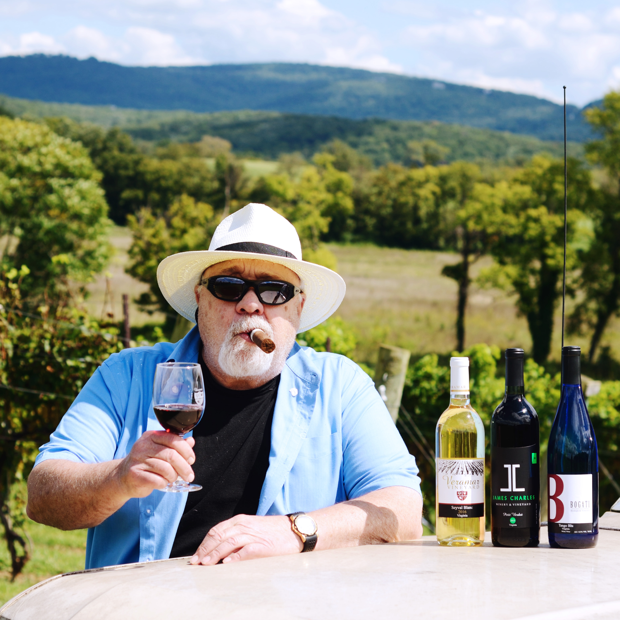 A man wearing a blue button down sits outside at the vineyard smoking a cigar and holding a glass of wine while 3 wine bottles sit next to him on the table on the home page.