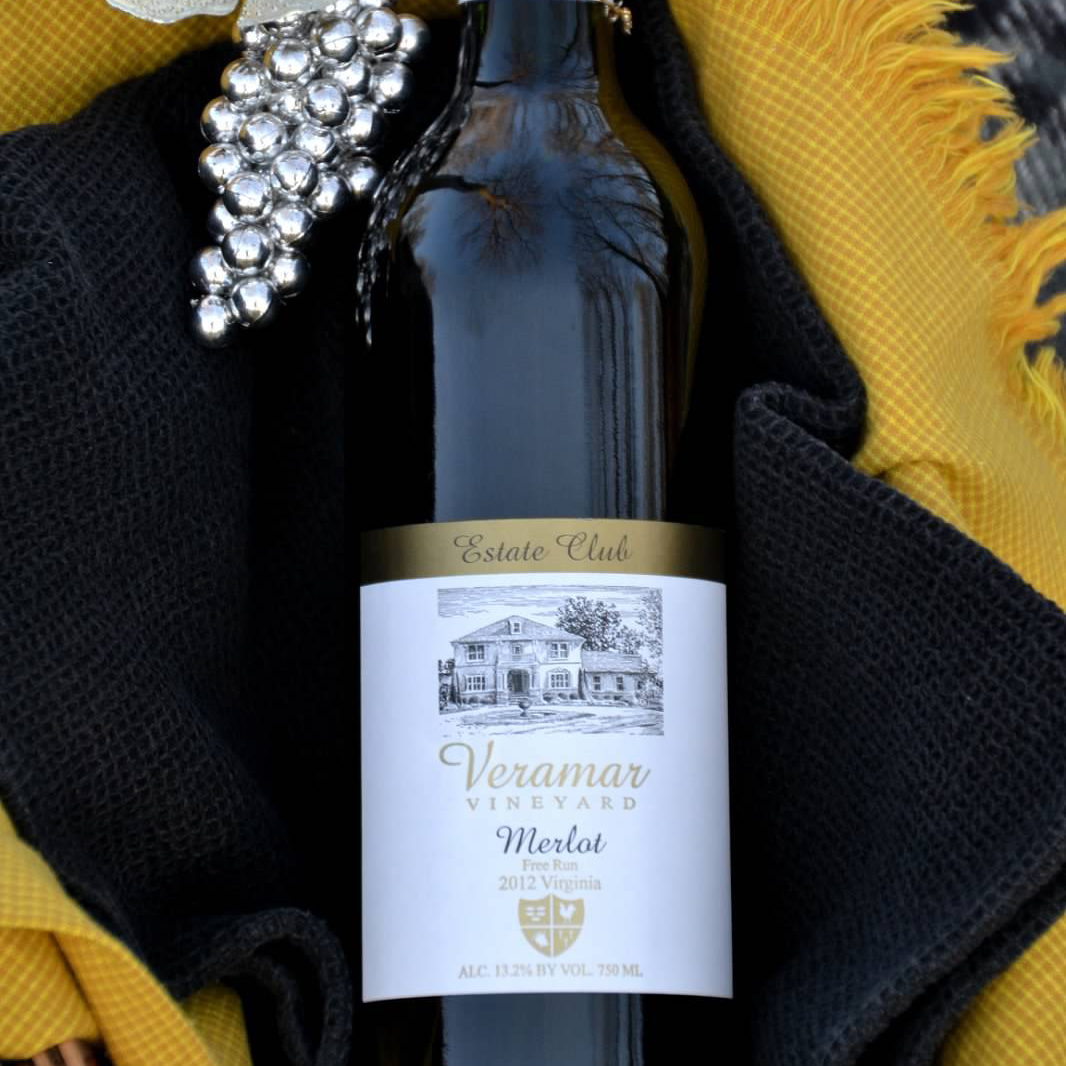 a wine bottle labeled Estate Club Veramar Vineyard Merlot lies on top of a black and yellow cloth with a silver decorative bunch of grapes next to it on the home page.