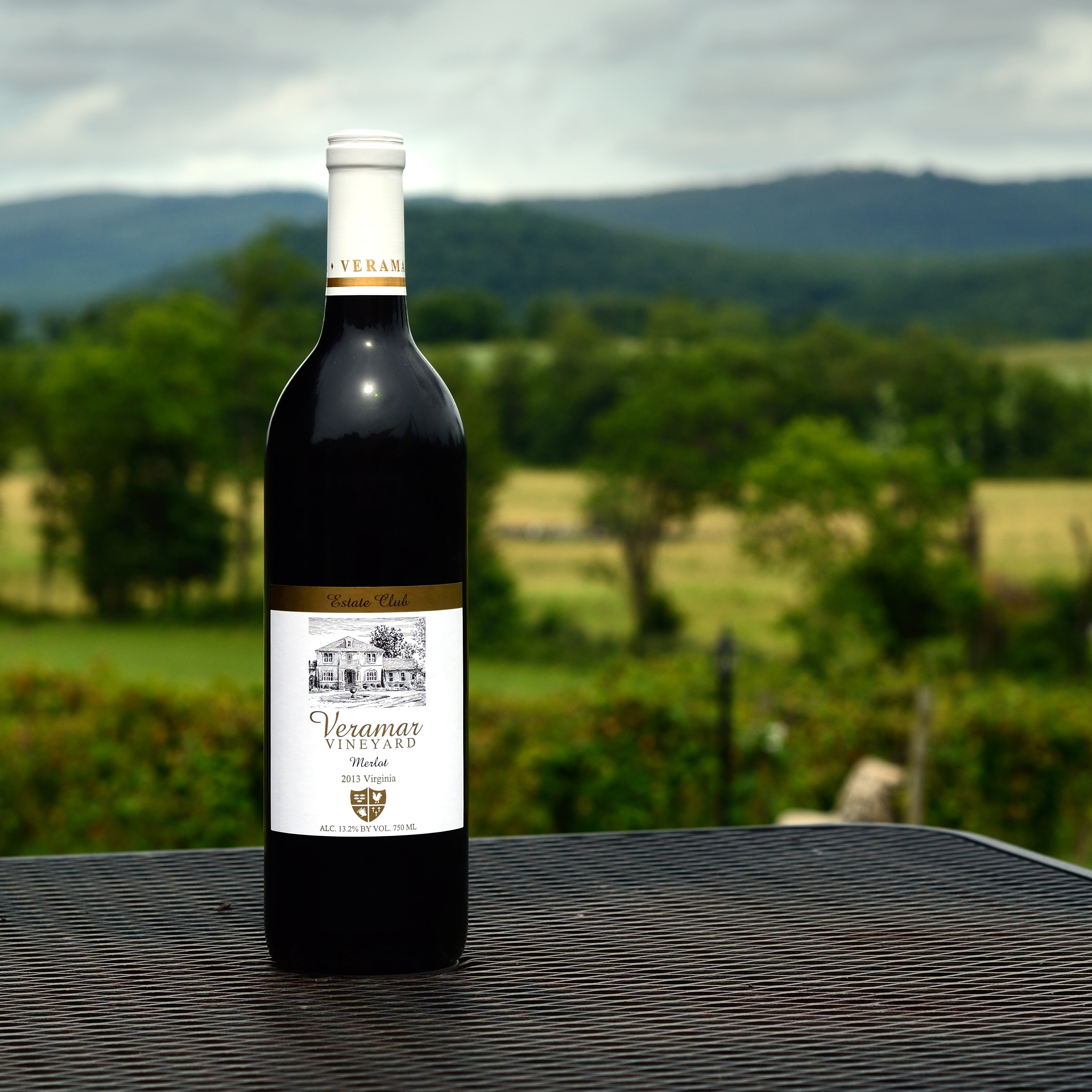 A bottle of Veramar Vineyard Merlot wine prominently displayed on a table with a scenic backdrop of rolling hills and lush greenery on the home page.